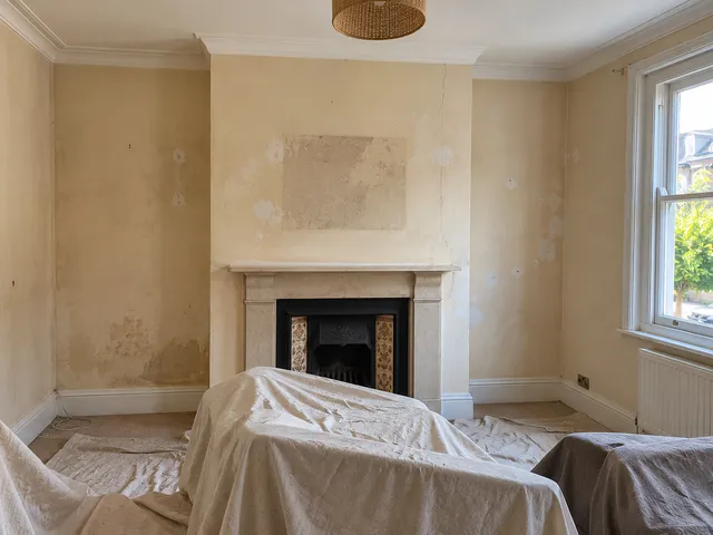 A living room with stained, patchy walls and furniture under dust sheets.