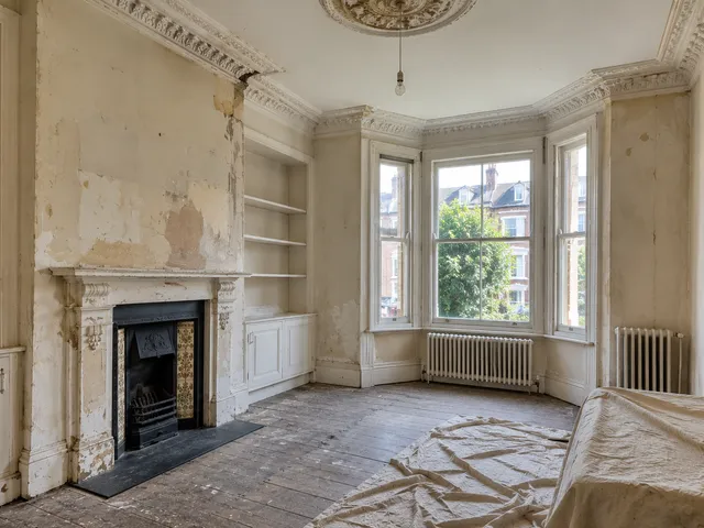 A period sitting room with a damaged fireplace surround, cracked cornicing and stripped walls.